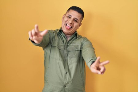 Téléchargez les photos : Jeune homme hispanique debout sur fond jaune souriant avec la langue montrant les doigts des deux mains faisant signe de victoire. numéro deux. - en image libre de droit