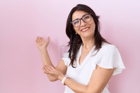 Foto de Mujer hispana de mediana edad vistiendo camiseta blanca casual y gafas invitando a entrar sonriendo natural con la mano abierta - Imagen libre de derechos