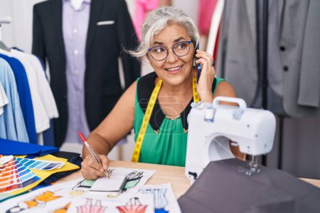 Foto de Middle age grey-haired woman tailor talking on smartphone drawing clothing design at tailor shop - Imagen libre de derechos