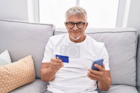Foto de Hombre de pelo gris de mediana edad con teléfono inteligente y tarjeta de crédito sentado en el sofá en casa - Imagen libre de derechos