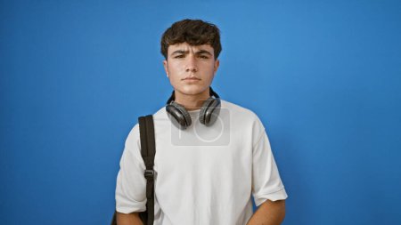 Photo for Cool young hispanic student guy wearing headphones and backpack, absorbed in his tech gadget. isolated portrait of a casual and smart teenager against a blue wall. - Royalty Free Image