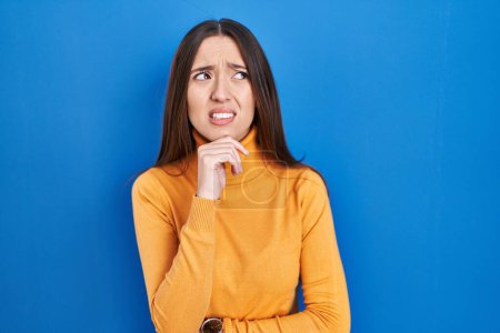 Photo for Young brunette woman standing over blue background thinking worried about a question, concerned and nervous with hand on chin - Royalty Free Image