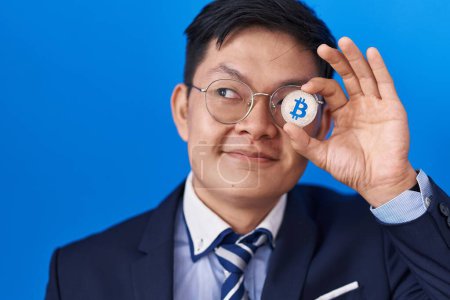 Foto de Joven asiático hombre celebración virtual moneda bitcoin cubriendo ojo sonriendo mirando a un lado y mirando hacia otro pensamiento. - Imagen libre de derechos