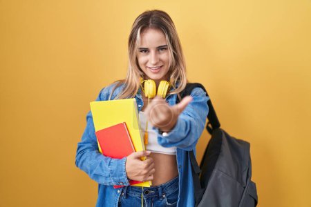 Foto de Joven mujer rubia vistiendo mochila de estudiante y sosteniendo libros haciendo señas ven aquí gesto con la mano invitando a la bienvenida feliz y sonriente - Imagen libre de derechos