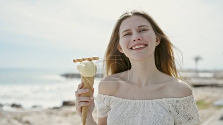 Foto de Joven mujer rubia turista sonriendo confiado sosteniendo helado en la playa - Imagen libre de derechos