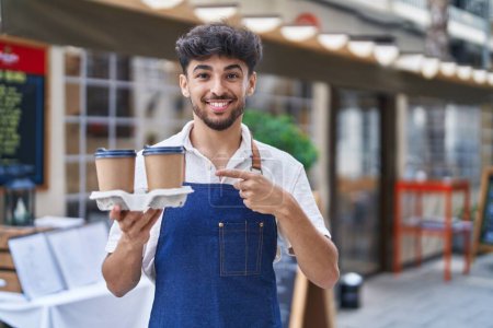 Téléchargez les photos : Arabe avec barbe portant tablier de serveur à la terrasse du restaurant souriant heureux pointant avec la main et le doigt - en image libre de droit