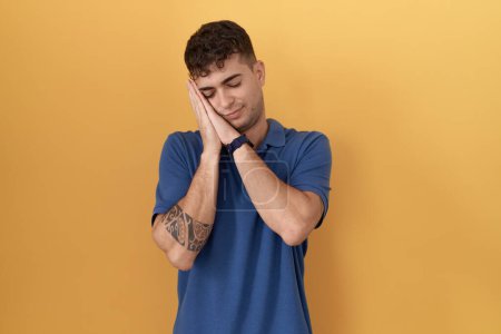 Photo for Young hispanic man standing over yellow background sleeping tired dreaming and posing with hands together while smiling with closed eyes. - Royalty Free Image