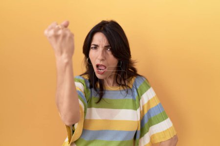 Foto de Mujer hispana de mediana edad de pie sobre fondo amarillo enojado y loco levantando el puño frustrado y furioso mientras gritaba con ira. rabia y concepto agresivo. - Imagen libre de derechos