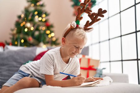 Foto de Adorable escritura infantil en el cuaderno acostado en el sofá por el árbol de Navidad en casa - Imagen libre de derechos