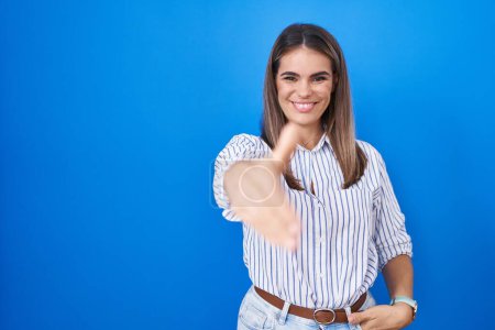Foto de Mujer joven hispana de pie sobre fondo azul sonriendo amistoso ofreciendo apretón de manos como saludo y bienvenida. negocios exitosos. - Imagen libre de derechos