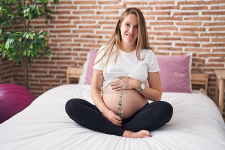 Téléchargez les photos : Jeune femme enceinte touchant le ventre tenant plante dans la chambre à coucher - en image libre de droit