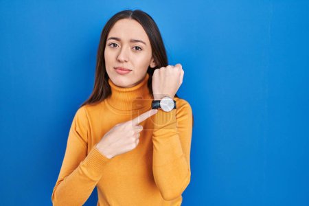 Photo for Young brunette woman standing over blue background in hurry pointing to watch time, impatience, looking at the camera with relaxed expression - Royalty Free Image
