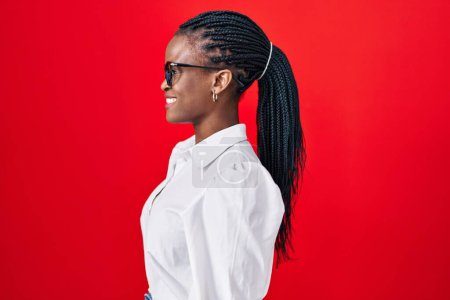 Téléchargez les photos : Femme africaine avec des tresses debout sur fond rouge regardant vers le côté, pose de profil relax avec visage naturel avec sourire confiant. - en image libre de droit