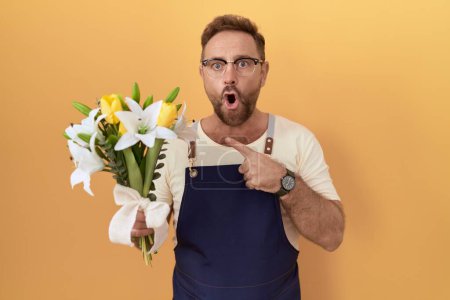 Photo for Middle age man with beard florist shop holding flowers surprised pointing with finger to the side, open mouth amazed expression. - Royalty Free Image