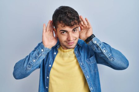 Téléchargez les photos : Jeune homme hispanique debout sur fond bleu essayant d'entendre les deux mains sur le geste de l'oreille, curieux de commérages. problème auditif, sourd - en image libre de droit
