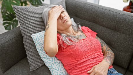Foto de Mujer estresada de pelo gris de mediana edad que yace sola en el sofá de su casa, consumida por la depresión, la frustración y la tristeza dolorosa. - Imagen libre de derechos