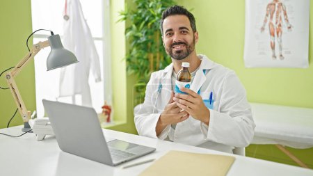 Téléchargez les photos : Jeune homme hispanique médecin en utilisant un ordinateur portable tenant bouteille de médicaments à la clinique - en image libre de droit