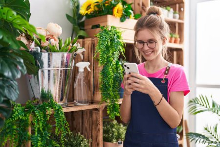 Téléchargez les photos : Jeune femme blonde fleuriste souriant confiant en utilisant un smartphone à la boutique de fleurs - en image libre de droit