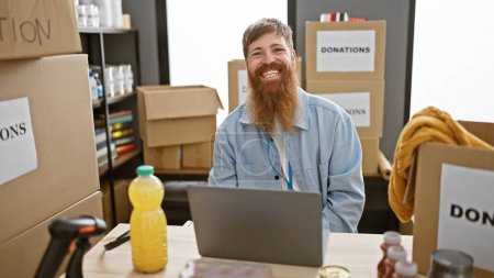 Photo for Happy, confident young redhead man sitting at a table, smiling, working on laptop indoors at a charity center, providing his service as a volunteer - Royalty Free Image