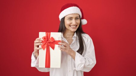 Foto de Joven hermosa mujer hispana sonriendo confiada sosteniendo regalo de Navidad sobre fondo rojo aislado - Imagen libre de derechos