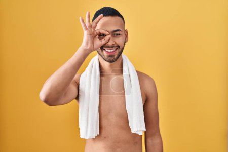 Foto de Joven hombre hispano de pie sin camisa con toalla haciendo buen gesto con la mano sonriente, los ojos mirando a través de los dedos con cara feliz. - Imagen libre de derechos