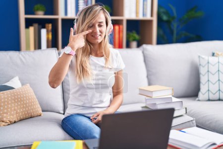Foto de Mujer rubia joven que estudia utilizando el ordenador portátil en casa señalando con la mano el dedo a la cara y la nariz, sonriendo alegre. concepto de belleza - Imagen libre de derechos