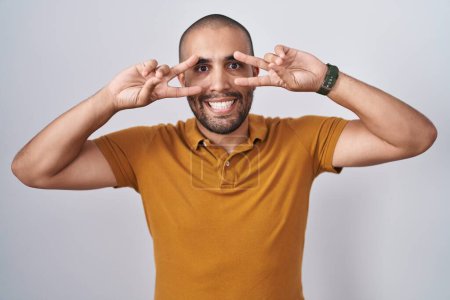 Téléchargez les photos : Hispanic man with beard standing over white background doing peace symbol with fingers over face, smiling cheerful showing victory - en image libre de droit