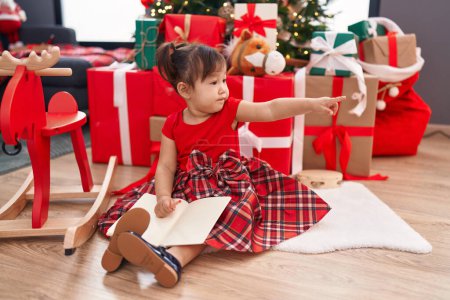 Foto de Adorable chica china señalando a un lado con el dedo sentado en el suelo por el árbol de Navidad en casa - Imagen libre de derechos
