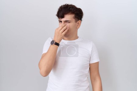 Téléchargez les photos : Jeune homme non binaire portant un t-shirt blanc décontracté sentant quelque chose de puant et dégoûtant, odeur intolérable, retenant son souffle avec les doigts sur le nez. mauvaise odeur - en image libre de droit