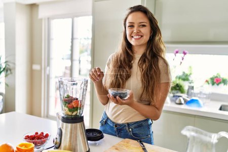 Photo for Young beautiful hispanic woman preparing vegetable smoothie with blender holding bowl with blueberries at the kitchen - Royalty Free Image