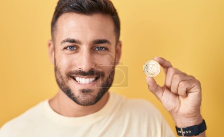 Foto de Hombre hispano guapo sosteniendo moneda criptomoneda tron mirando positiva y feliz de pie y sonriendo con una sonrisa confiada mostrando los dientes - Imagen libre de derechos