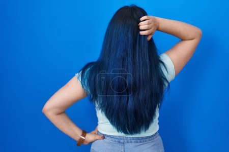 Téléchargez les photos : Jeune fille moderne avec les cheveux bleus debout sur fond bleu vers l'arrière en pensant au doute avec la main sur la tête - en image libre de droit