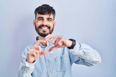 Photo for Young hispanic man with beard standing over blue background smiling in love doing heart symbol shape with hands. romantic concept. - Royalty Free Image
