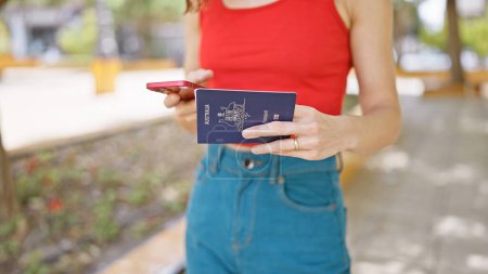 Téléchargez les photos : Les mains d'une femme américaine indépendante tenant son passeport à l'extérieur, tapant avec désinvolture un message sur son smartphone, se promenant dans un parc urbain animé avant son passionnant voyage de vacances - en image libre de droit