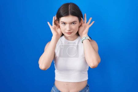 Foto de Mujer caucásica joven de pie sobre fondo azul tratando de escuchar ambas manos en el gesto de la oreja, curioso por chismes. problema auditivo, sordo - Imagen libre de derechos