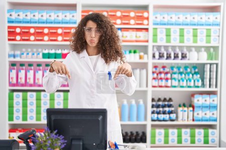 Téléchargez les photos : Femme hispanique avec les cheveux bouclés travaillant à la pharmacie pharmacie pointant vers le bas regardant triste et contrarié, indiquant la direction avec les doigts, malheureux et déprimé. - en image libre de droit