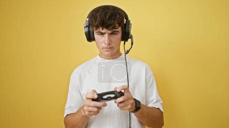 Photo for Upset young hispanic teenager, engrossed in playing a video game, stands isolated against a yellow background expressing anger, possibly losing - Royalty Free Image