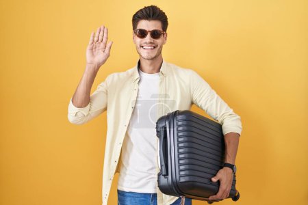 Téléchargez les photos : Jeune homme hispanique tenant une valise allant en vacances d'été renonçant à dire bonjour heureux et souriant, geste de bienvenue amical - en image libre de droit