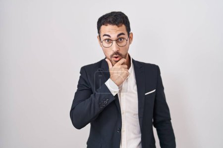 Photo for Handsome business hispanic man standing over white background looking fascinated with disbelief, surprise and amazed expression with hands on chin - Royalty Free Image