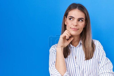 Photo for Hispanic young woman standing over blue background with hand on chin thinking about question, pensive expression. smiling with thoughtful face. doubt concept. - Royalty Free Image