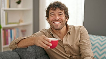 Téléchargez les photos : Jeune homme hispanique tenant une tasse de café assis sur un canapé souriant à la maison - en image libre de droit