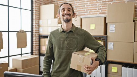 Foto de Joven hispano hombre comercio electrónico trabajador de negocios sosteniendo paquete sonriendo en la oficina - Imagen libre de derechos