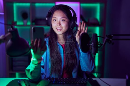 Foto de Joven mujer asiática jugando videojuegos con smartphone renunciando a decir hola feliz y sonriente, gesto de bienvenida amistoso - Imagen libre de derechos