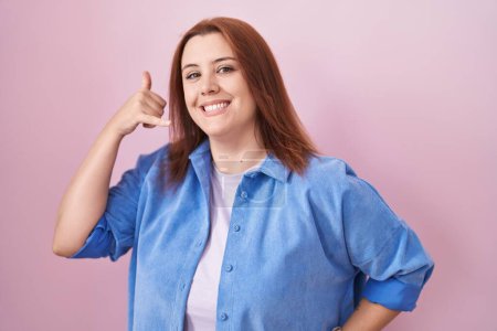 Foto de Mujer hispana joven con el pelo rojo de pie sobre fondo rosa sonriendo haciendo gesto de teléfono con la mano y los dedos como hablar por teléfono. comunicar conceptos. - Imagen libre de derechos