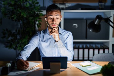 Photo for Young hispanic man working at the office at night thinking concentrated about doubt with finger on chin and looking up wondering - Royalty Free Image
