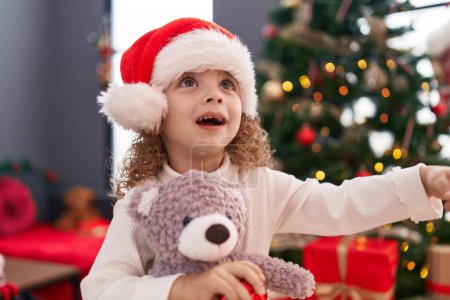 Photo for Adorable blonde girl hugging teddy bear standing by christmas tree at home - Royalty Free Image
