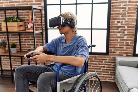 Foto de Joven hombre rubio jugando videojuego con gafas de realidad virtual y joystick sentado en silla de ruedas en casa - Imagen libre de derechos