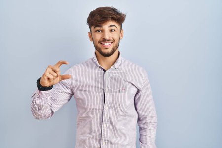 Téléchargez les photos : Homme arabe avec barbe debout sur fond bleu geste souriant et confiant avec la main faisant signe de petite taille avec les doigts regardant et la caméra. concept de mesure. - en image libre de droit