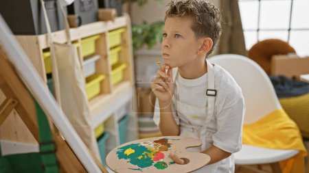 Téléchargez les photos : Adorable garçon artiste blond, dessinant avec confiance dans un studio d'art intérieur, regardant la toile avec réflexion, tablier et pinceau à la main. - en image libre de droit