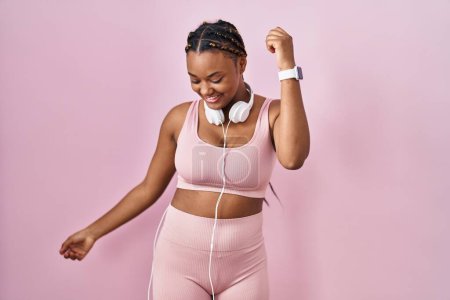 Téléchargez les photos : Femme afro-américaine avec des tresses portant des vêtements de sport et des écouteurs dansant heureux et joyeux, souriant décontracté et confiant écoutant de la musique - en image libre de droit